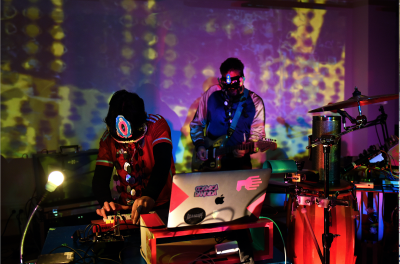 Zu sehen ist ein DJ, der das Mischpult bedient, und ein Gitarist, beide werden von bunten Lichtern angeleuchtet.