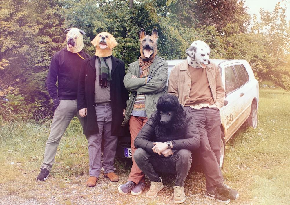 Gruppenfoto mit 5 Menschen mit Hundeköpfen und einem alten Auto im Hintergrund